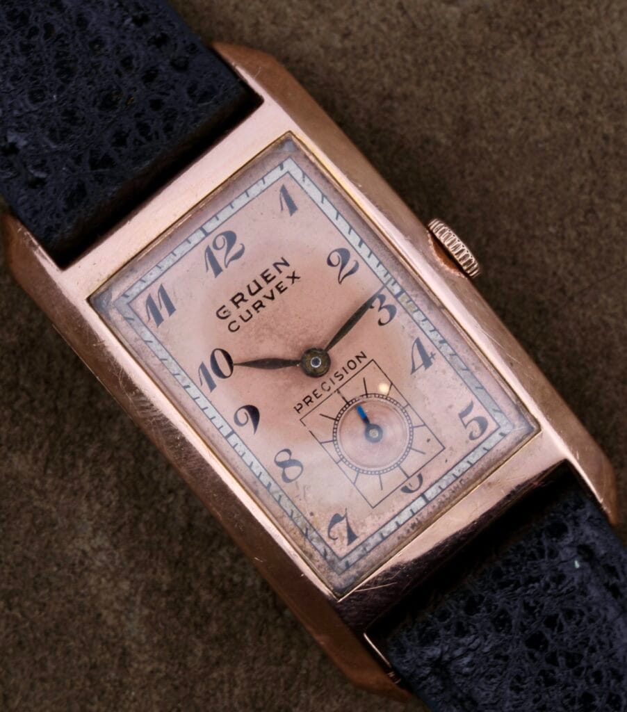 A vintage Gruen Curvex Salmon Dial 14k Rose Gold wristwatch with a black textured strap, displayed on a brown surface.