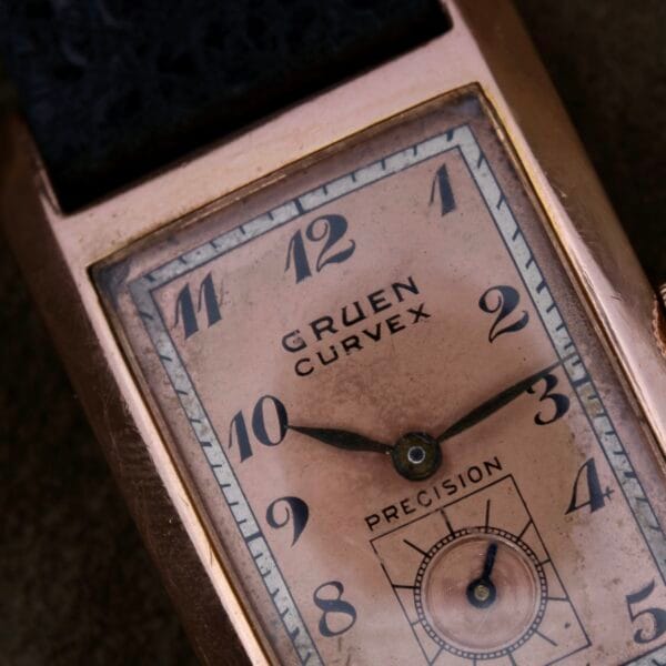 Close-up of a Gruen Curvex Salmon Dial wristwatch in 14k rose gold, with a rectangular curved face. It has black numerals, minute markers, a small seconds subdial, and hands at 10:10 within its casing, paired with a sleek black strap.