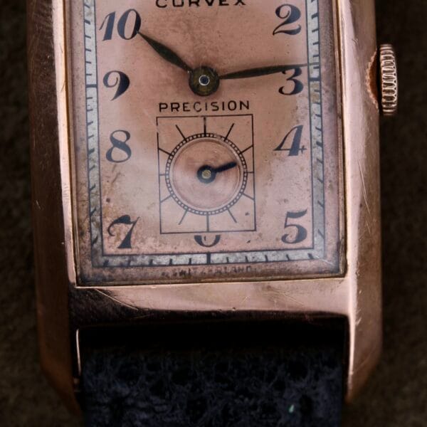 Close-up of a vintage Gruen Curvex Salmon Dial wristwatch with a 14k rose gold case. The elegant salmon dial features numbers 1 to 12 and an additional seconds dial. Its black textured leather strap displays wear, enhancing its antique charm.