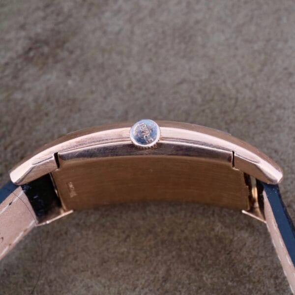 Close-up image of the Gruen Curvex Salmon Dial 14k Rose Gold wristwatch on its side, showcasing the logo-engraved crown. The salmon dial adds elegance as the watch band rests on a textured brown surface featuring intricate crown detailing.