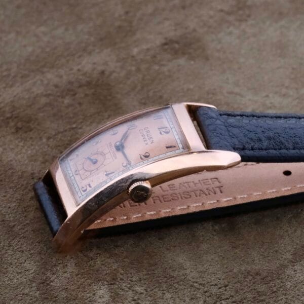 Close-up of a vintage Gruen Curvex Salmon Dial 14k Rose Gold wristwatch, featuring a rectangular gold-toned case, black leather strap, refined salmon dial with black Arabic numerals, and a small seconds subdial at 6 oclock on a textured brown surface.