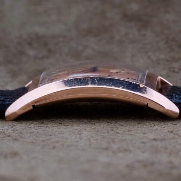 A close-up of a Gruen Curvex Salmon Dial wristwatch showcases its curved 14k rose gold case with a scratched crystal. Although the salmon dial remains unseen, the dark strap rests elegantly on a textured gray surface, reminiscent of vintage charm.