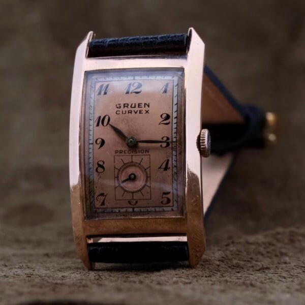 The Gruen Curvex Salmon Dial wristwatch, in 14k rose gold, boasts a rectangular case and black leather strap. Its unique salmon dial with Arabic numerals and a small seconds subdial elegantly stands on a textured brown surface.