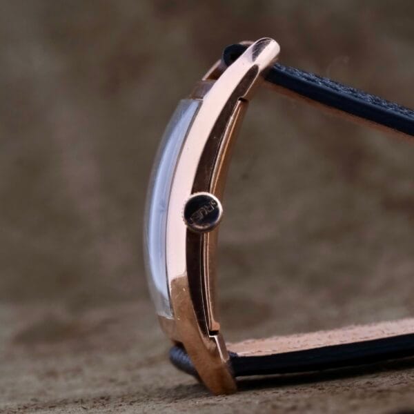 A close-up side view of the Gruen Curvex Salmon Dial 14k Rose Gold wristwatch showcases a gold-toned case, black leather strap, salmon dial, protruding crown, and slightly curved crystal against a blurred brown background.
