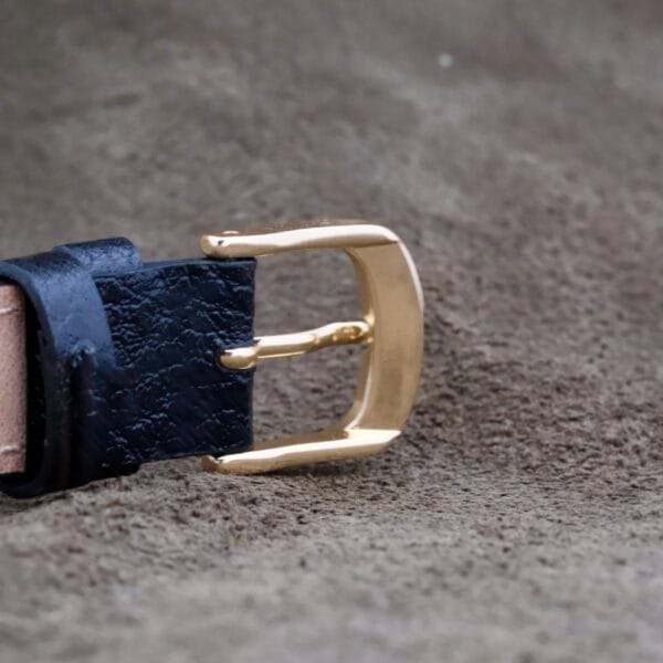 Close-up of a gold-colored belt buckle, echoing the elegance of the Gruen Curvex Salmon Dial 14k Rose Gold, attached to a black leather strap and resting on a textured brown surface.
