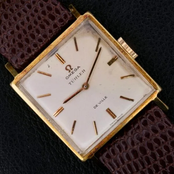A vintage Omega Deville Turler features a square gold case, an off-white dial, and a brown leather strap. It has minimalist hour markers and hands with the brand and model elegantly displayed on the dial.