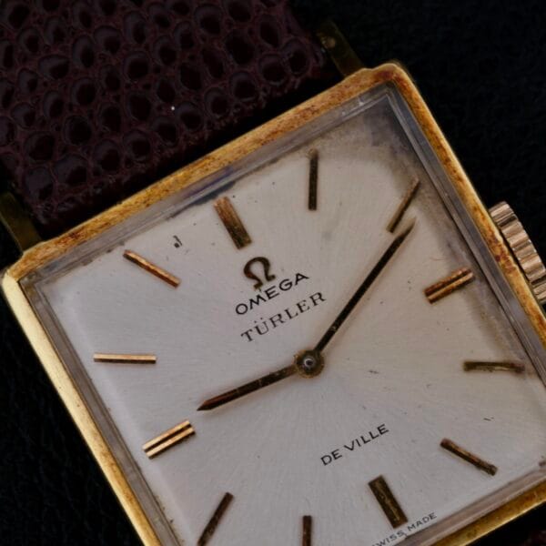 Close-up of an Omega Deville Turler with a square gold case and brown leather strap, showcasing a minimalist design with gold hour markers on its elegant dial. The precision hands move over the white face, marked Swiss Made at the bottom.