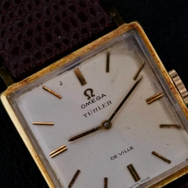 Close-up of an Omega Deville Turler with a square gold case and brown leather strap, showcasing a minimalist design with gold hour markers on its elegant dial. The precision hands move over the white face, marked Swiss Made at the bottom.