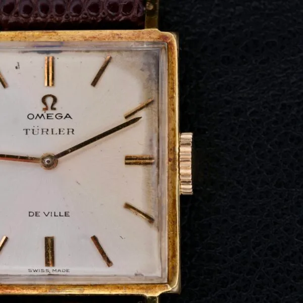 Close-up of a vintage Omega Deville Turler Dial Square Case wristwatch, featuring gold hour markers, two hands, and Omega, TÜRLER, and De Ville inscriptions on its dial. The rectangular gold-tone case is paired with an elegant brown leather strap.
