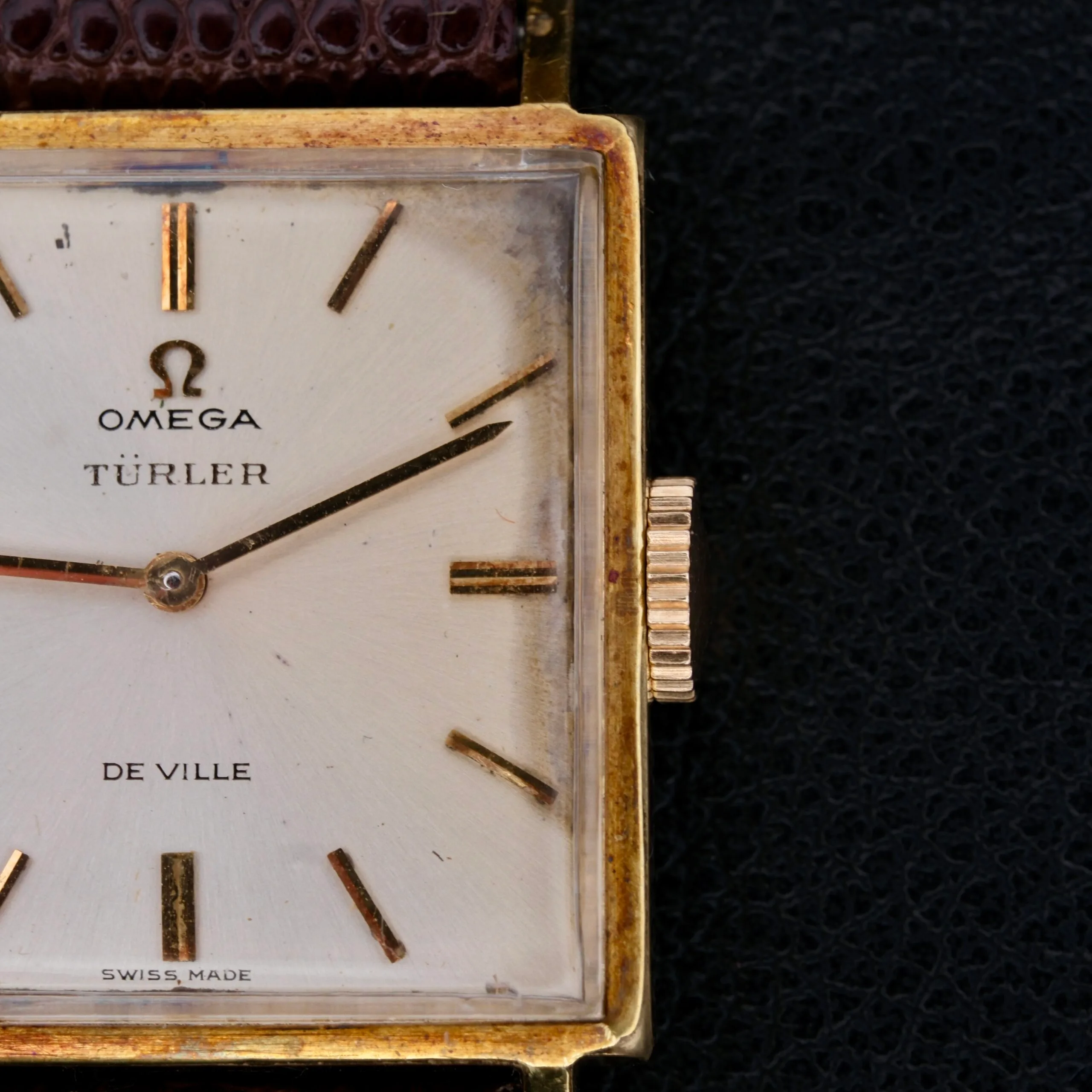 Close-up of a vintage Omega Deville Turler Dial Square Case wristwatch, featuring gold hour markers, two hands, and Omega, TÜRLER, and De Ville inscriptions on its dial. The rectangular gold-tone case is paired with an elegant brown leather strap.