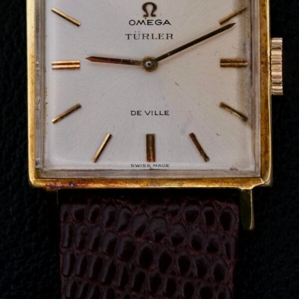The Omega Deville Turler Dial Square Case watch showcases a vintage square gold design with a unique Türler dial and minimalist gold hour markers. Its textured brown leather strap adds elegance, and Swiss Made is inscribed at the bottom.