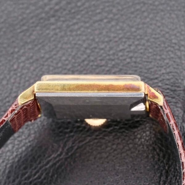 The Omega Deville Turler Dial Square Case wristwatch, featuring a rectangular face, gold edges, and a dark strap, rests face down on a textured black surface.