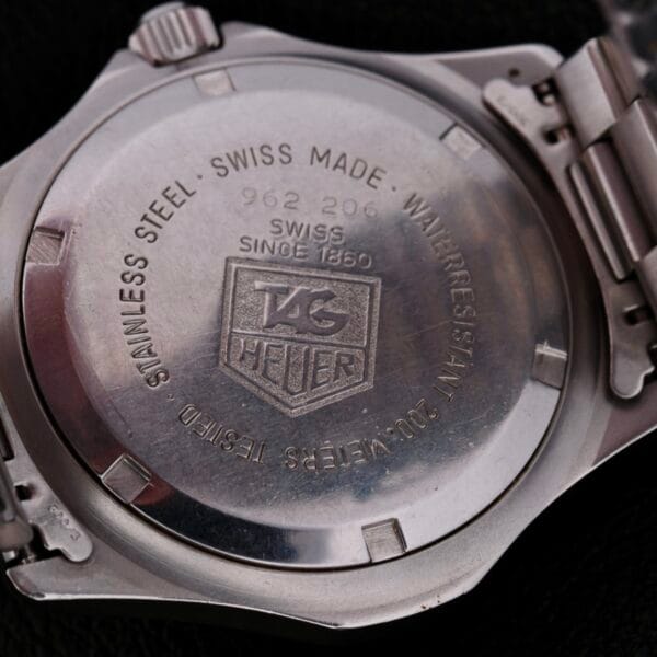 Close-up of the Tag Heuer Professional 2000 Dive watch back, displaying engravings like Swiss Made, Water Resistant 200 Meters, the TAG Heuer logo, and Stainless Steel. The watch features a sleek metal bracelet and crown resting on a leather surface.