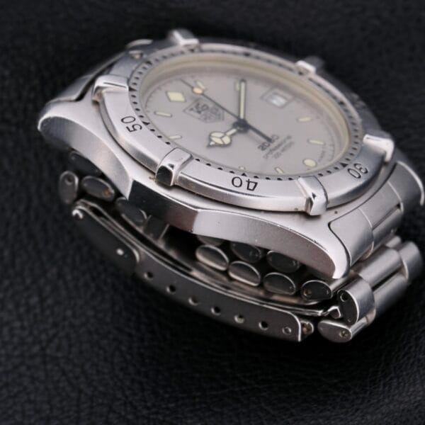 A close-up of the Tag Heuer Professional 2000 Dive wristwatch highlights its silver metal band and round face with time markers and a date display. Set on a black leather surface, this elegant timepiece exudes sophistication and precision.