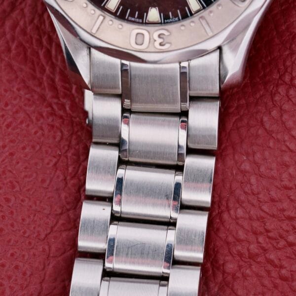 Close-up of a stainless steel Omega Seamaster Professional Chronometer bracelet on textured red leather. The partially visible watch face reveals part of the bezel with markings, including 30. The bracelet features a brushed metal finish and secure clasp.
