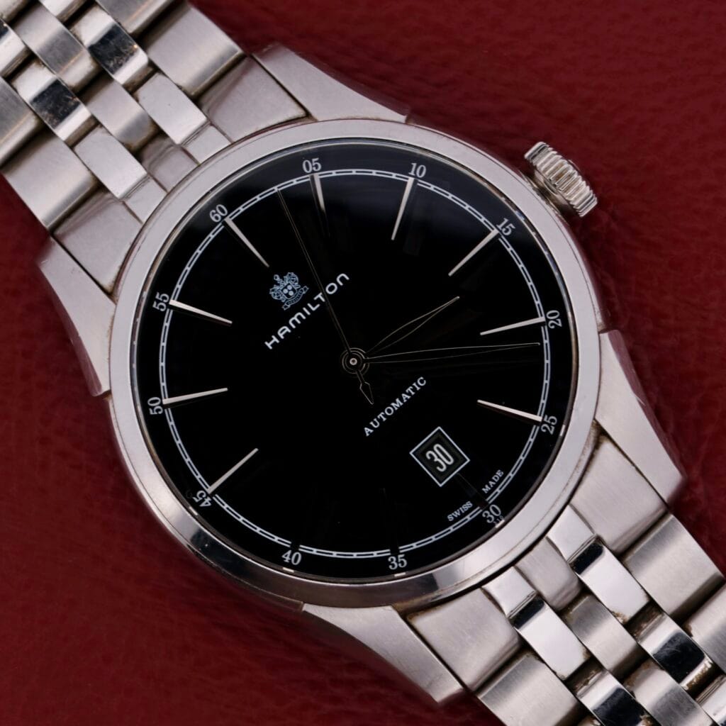 Hamilton American Classic Spirit Of Liberty watch with a black dial and metal band, displayed on a textured red surface.