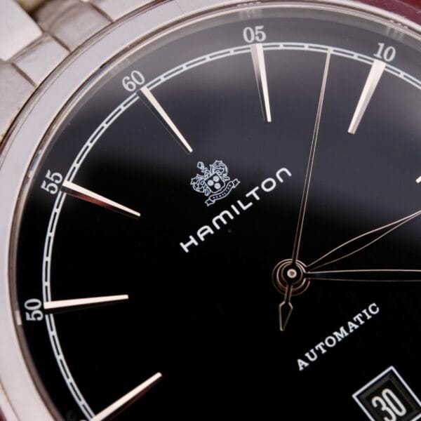 The Hamilton American Classic Spirit of Liberty wristwatch features a black dial with silver hour markers, the Hamilton logo with crest, Automatic, and a date display at the bottom right. It includes a metal bezel and band.