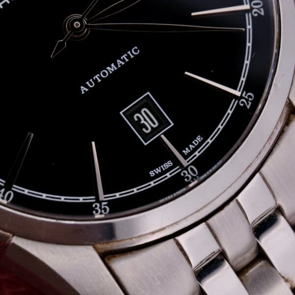 Close-up of the Hamilton American Classic Spirit Of Liberty watch face, featuring a black dial with silver hour markers and hands. The date 30 is shown in a box. Automatic and Swiss Made are visible on this luxury timepiece, which includes a stainless steel band.