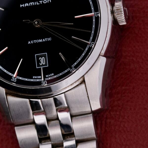 Close-up of a Hamilton American Classic Spirit Of Liberty watch with an automatic movement and stainless steel band. Its sleek black dial, featuring slim metallic hands and a date window displaying 30, rests elegantly on a textured red leather surface.