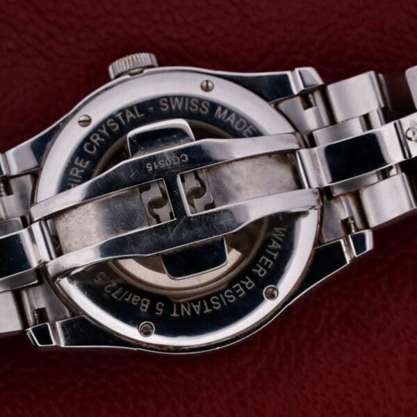 The image showcases the back of a Swiss-made Hamilton American Classic Spirit Of Liberty watch with a metal bracelet, engraved with Sapphire Crystal and Water Resistant 5 Bar, resting elegantly against a textured dark red surface.