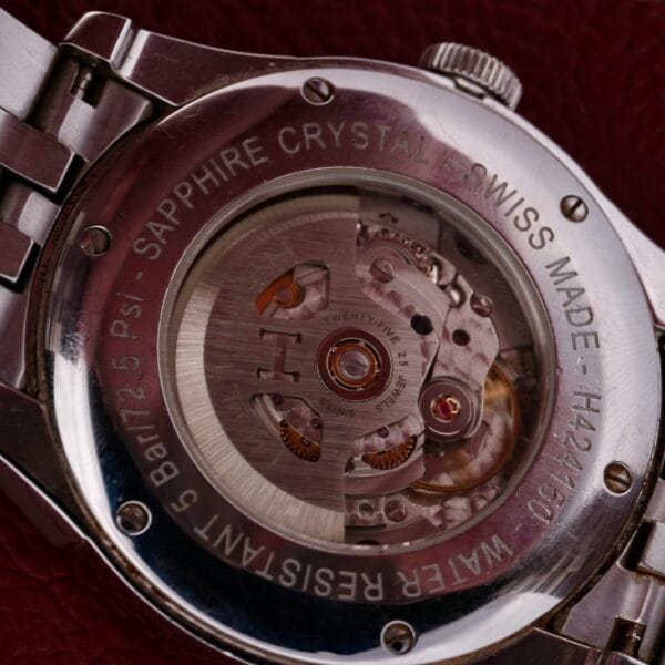 A close-up of the Hamilton American Classic Spirit Of Libertys back reveals a transparent case showcasing intricate mechanical movement, with details about sapphire crystal, Swiss manufacturing, water resistance, and model number engraved on the metal. The watch rests elegantly on leather.