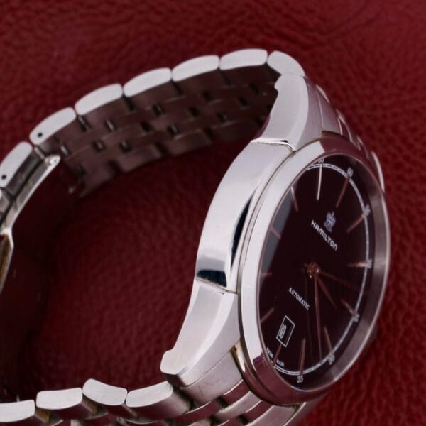The Hamilton American Classic Spirit Of Liberty features a silver case, dark brown face with a date display, and Automatic inscribed. Elegantly positioned on a textured red surface, it highlights its sleek metal link strap.
