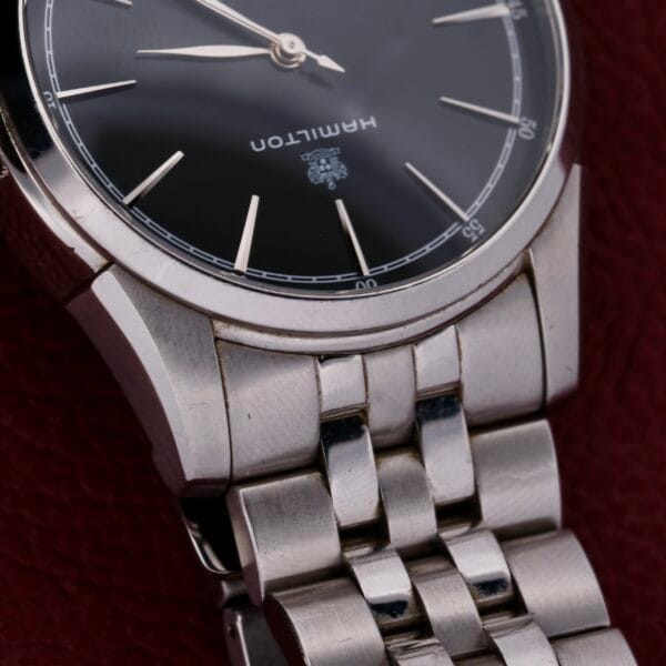 Close-up of the Hamilton American Classic Spirit Of Liberty wristwatch in silver with a dark dial and Hamilton branding. Resting on a dark red surface, it shows 10:09 and features an elegant metal link bracelet.