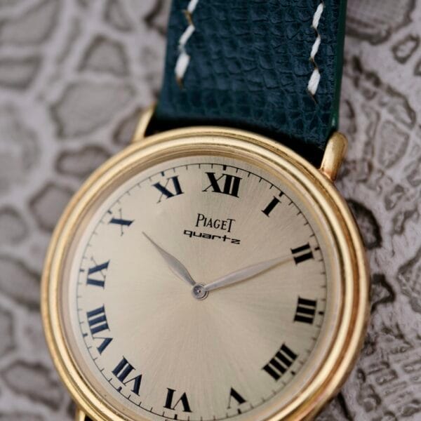 A close-up of the Piaget Quartz 7632 18k Gold Champagne Dial showcases its gold-toned round face with black Roman numerals and two elegant hands. The watch features a dark textured leather strap and is elegantly presented on a patterned surface.