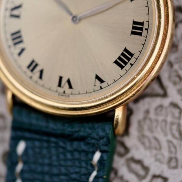 Close-up of a vintage Piaget Quartz 7632 18k Gold Champagne Dial watch, featuring a gold case, light face with black Roman numerals, and a textured dark green leather strap with visible stitching, all on a patterned fabric backdrop.