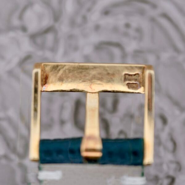 A close-up of a Piaget Quartz 7632 18k Gold watch buckle with engraved markings, elegantly positioned on a blue strap against a blurred gray backdrop.