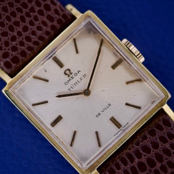 Omega Deville 111.071 with a rare Turler Dial, square gold case, and brown textured strap, displayed on a blue background.