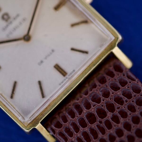 Close-up of an Omega Deville 111.071 Turler Dial Square Case with a white face and brown textured leather strap, set against a blue background.