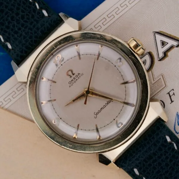 A close-up shows an Omega Seamaster Bull Horn Lug watch with a silver-tone dial, gold hour markers, and a black leather strap elegantly resting on paper featuring intricate blue and black designs.