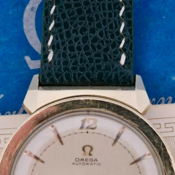 Detailed view of an Omega Seamaster Bull Horn Lug watch, featuring a textured dark strap and unique lugs. The dial displays the Omega logo and minimalist hour markers. The backdrop is a blue surface with partially visible white text.