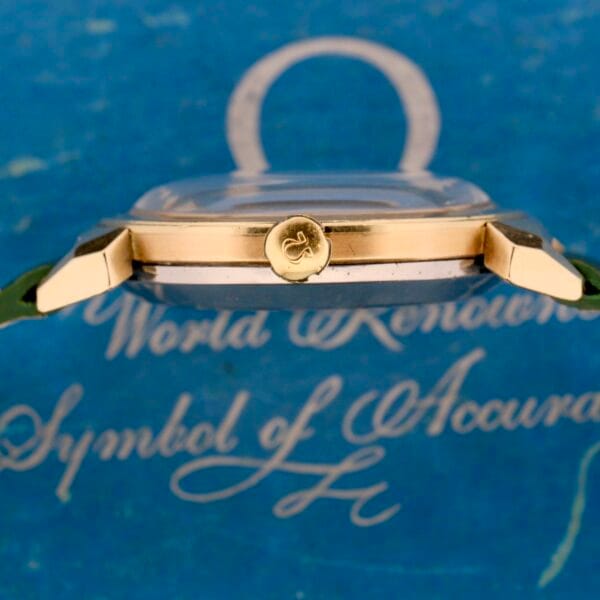 A side view of an Omega Seamaster Bull Horn Lug wristwatch showcases its gold crown and bull horn design. Set against a blue background, the watch bears white cursive text reading World Renowned Symbol of Accuracy and features a green strap with a transparent dome over the face.