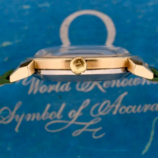 A side view of an Omega Seamaster Bull Horn Lug wristwatch showcases its gold crown and bull horn design. Set against a blue background, the watch bears white cursive text reading World Renowned Symbol of Accuracy and features a green strap with a transparent dome over the face.