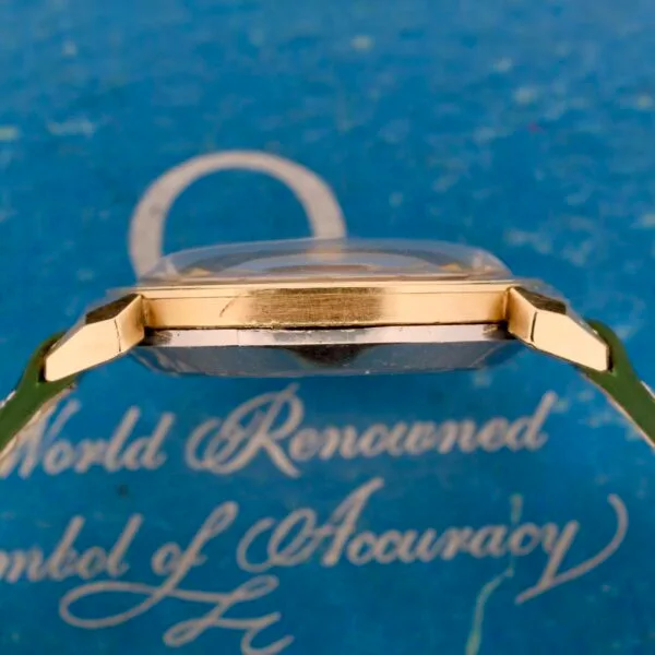 A close-up side view of the Omega Seamaster Bull Horn Lug wristwatch with a gold casing against a blue textured background. The background has partially visible white cursive text that reads World Renowned Symbol of Accuracy.