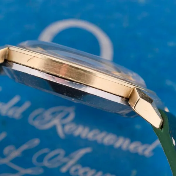Side view of the Omega Seamaster Bull Horn Lug with a gold case and green strap, set against a blurred blue background with white cursive writing. This watch features transparent crystal and shiny metal accents, highlighting its famous bull horn design.