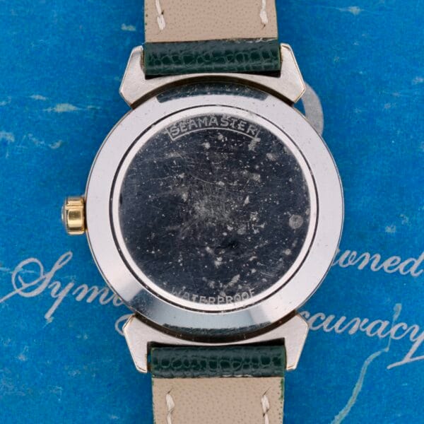 The image highlights the back of a vintage Omega Seamaster Bull Horn Lug watch. Despite scratches, its engraving is clear. It has a green leather strap and rests on a blue textured background with cursive writing, evoking the charm of its era.
