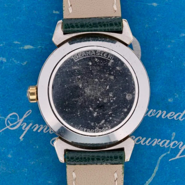 The image highlights the back of a vintage Omega Seamaster Bull Horn Lug watch. Despite scratches, its engraving is clear. It has a green leather strap and rests on a blue textured background with cursive writing, evoking the charm of its era.