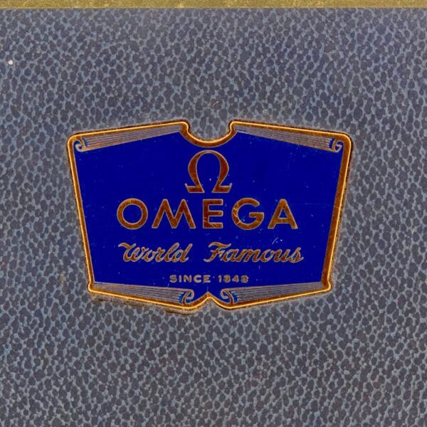 Close-up of a vintage Omega logo on a textured dark background. The design features the Omega Seamaster Bull Horn Lug style with elegant contours, reminiscent of classic craftsmanship and sophistication.