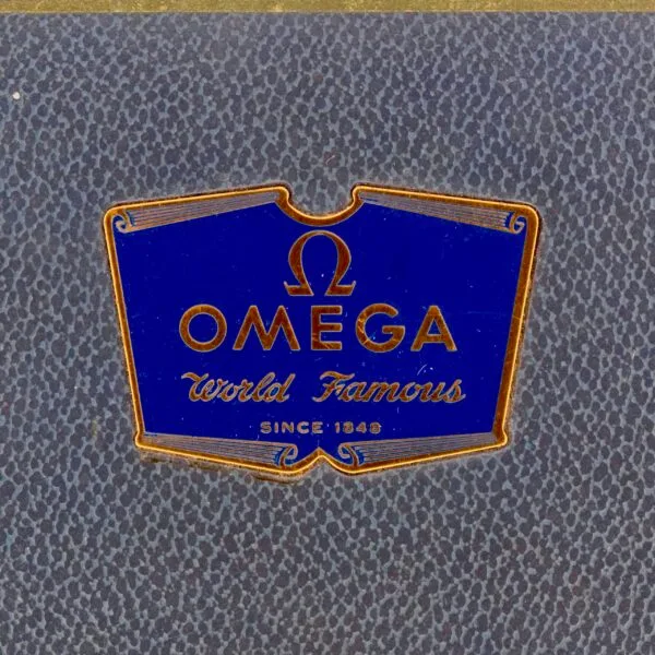 Close-up of a vintage Omega logo on a textured dark background. The design features the Omega Seamaster Bull Horn Lug style with elegant contours, reminiscent of classic craftsmanship and sophistication.