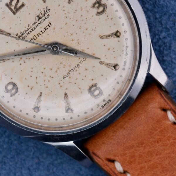 The IWC Automatic Turler Dial C. 852 features a vintage white speckled dial with silver hour markers, displaying International Watch Co. Schaffhausen and Automatic. It has a brown leather strap, beautifully set against a blue textured background.
