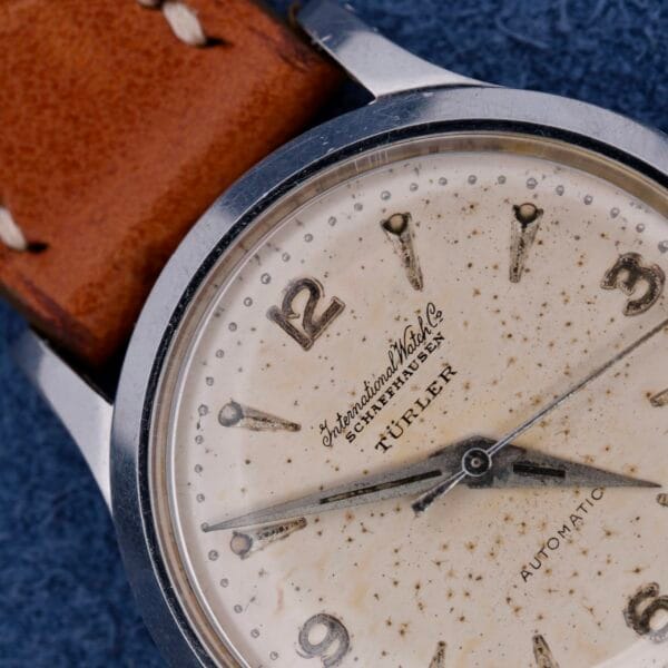 Close-up of the vintage IWC Automatic Turler Dial C. 852 wristwatch with a leather strap. The cream-colored dial shows aging, Arabic numerals, three hands, and inscriptions International Watch Co. Schaffhausen and Automatic above and below the center.