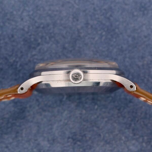 Side view of an IWC Automatic Turler Dial C. 852 wristwatch with a silver case, visible crown, and tan leather strap against a textured blue background.