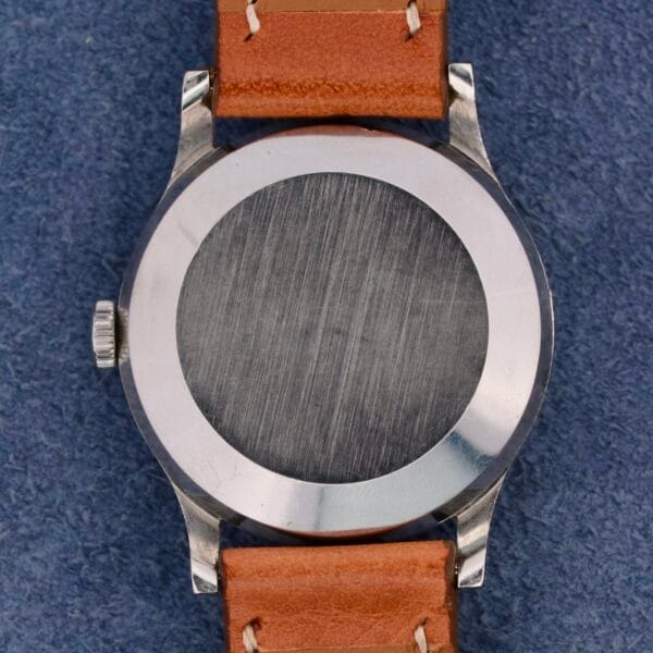 The image showcases the back of an IWC Automatic Turler Dial C. 852 wristwatch, featuring a round metal case with a brushed finish and a left-side crown. This automatic timepiece has a brown leather strap set against a textured blue fabric background.