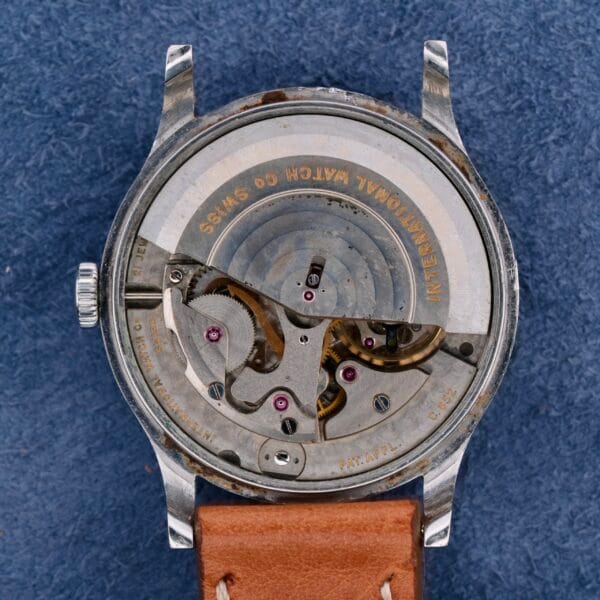 The image shows the back of an IWC Automatic Turler Dial C. 852 wristwatch, featuring an open case back with intricate gears engraved INTERNATIONAL WATCH CO. SWISS, set against a blue backdrop, complemented by a brown strap.