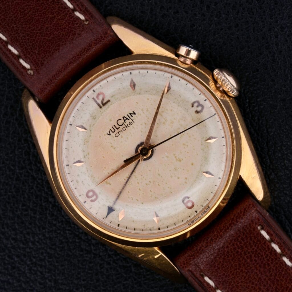 The Vulcain Cricket 17 Jewels watch with a brown leather strap rests on a black textured surface.