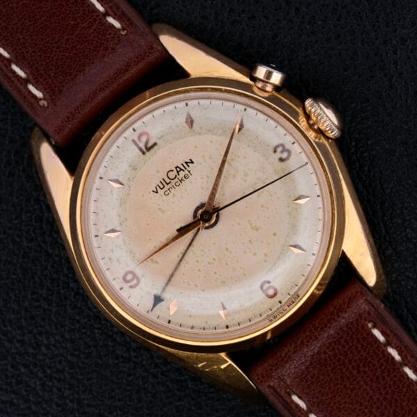 A vintage Vulcain Cricket 17 Jewels wristwatch featuring a gold casing, cream dial, and brown leather strap. It displays Arabic numerals at 9, 12, and 3 with gold hour markers and hands on a dark textured background.