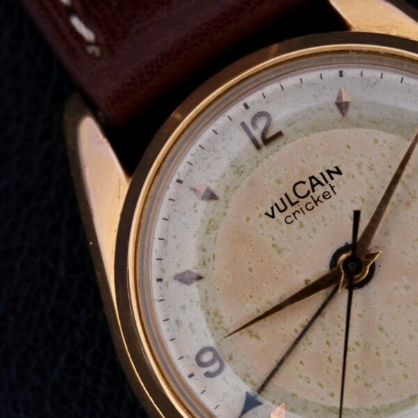 Close-up of a Vulcain Cricket 17 Jewels wristwatch with a gold-toned case, weathered cream dial featuring numbered hour markers, and pointed hands. Partially visible is its brown leather strap.